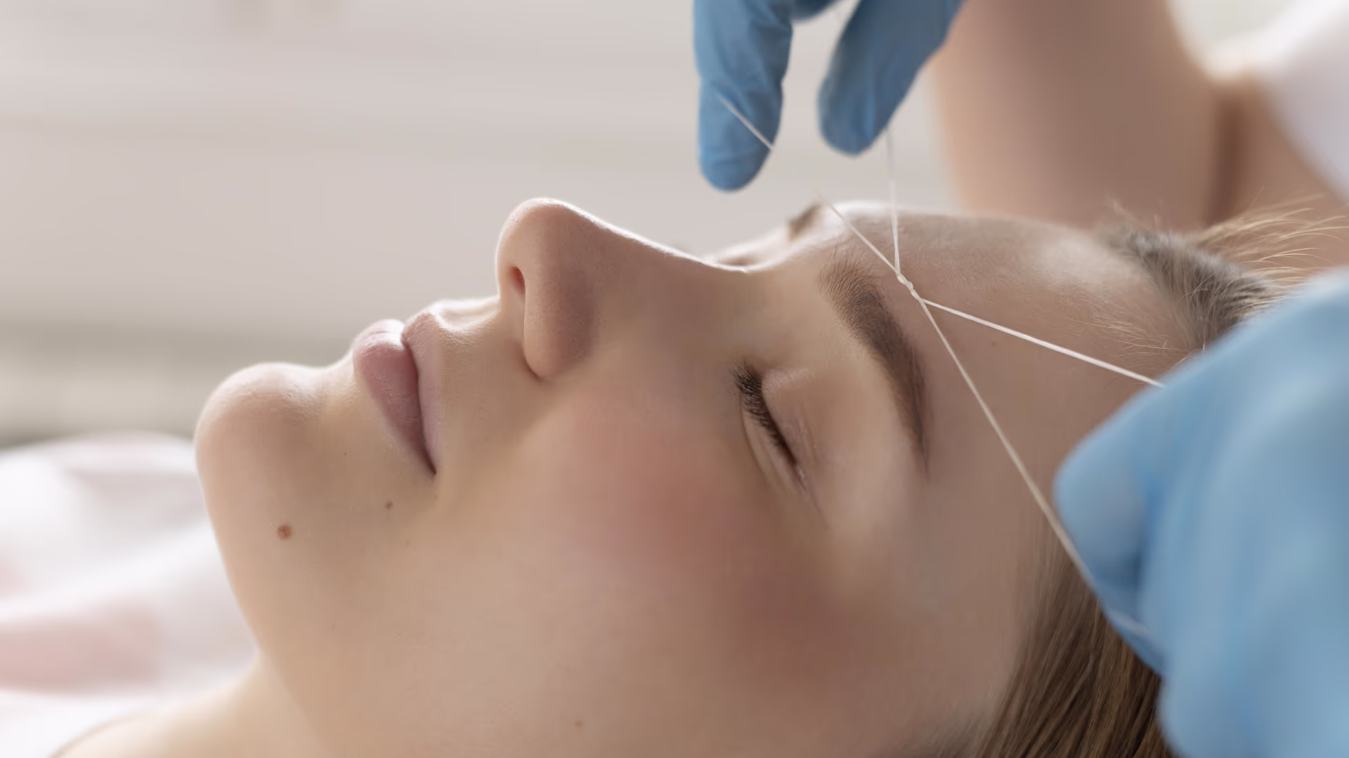 Profissional realizando depilação facial com linha na região da sobrancelha de cliente deitada